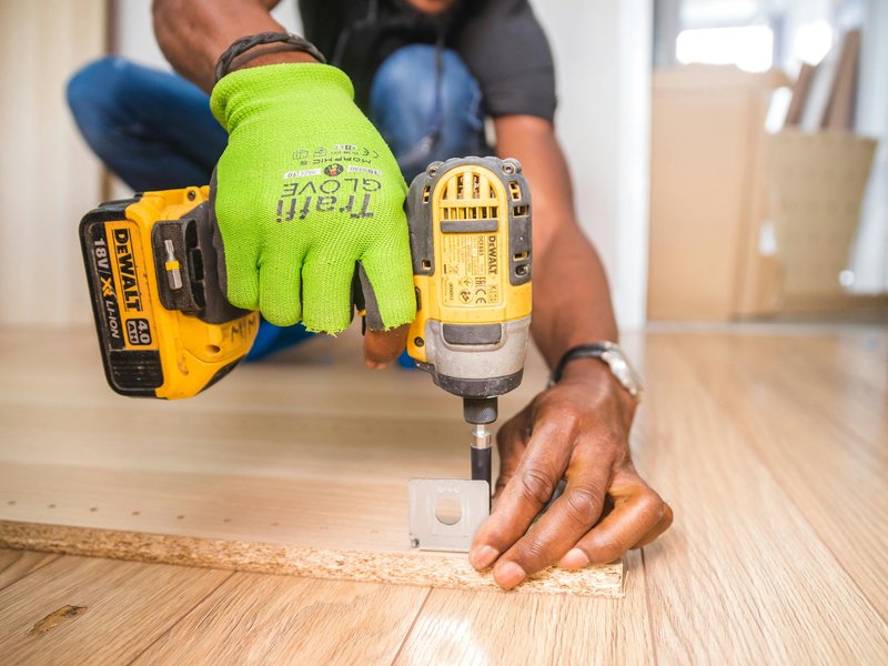 Close-up of a person assembling furniture or working on a DIY project, using a yellow DEWALT cordless drill to drive a screw into a particle board.