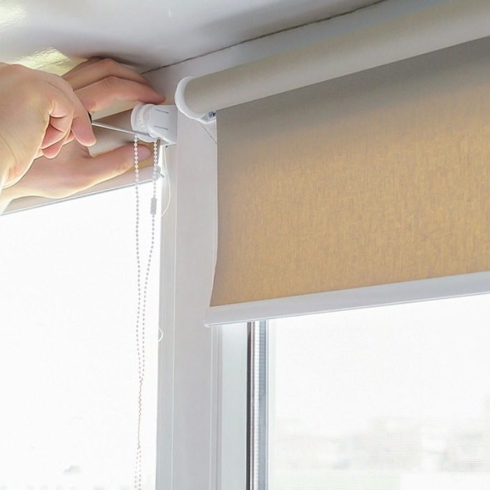 Close-up of a person adjusting a blackout roller blind inside a modern room, handling the chain mechanism at the top corner of the window.