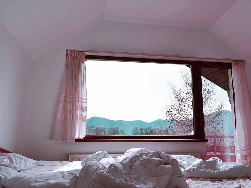 Cozy bedroom with an unmade bed featuring crumpled white sheets, set beneath a large window framed by pink curtains. Outside, tree branches and distant mountains are visible under a pale sky, creating a serene contrast between the peaceful interior and th